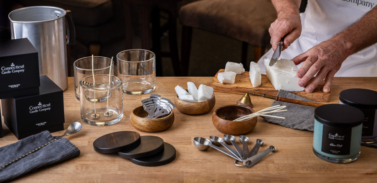 A person's hands are shown cutting a large white block of solid wax or soap on a wooden board, with several finished wax pieces nearby. In the foreground, a candle jar labeled 'Coconut Clay Candle Company' and measuring spoons are visible on a wooden table.

