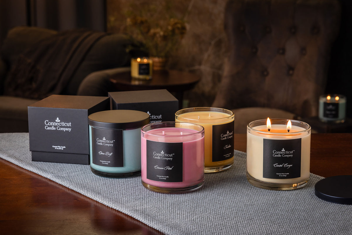 Four Connecticut Candle Company scented candles in glass jars with black labels, shown with their black gift boxes on a gray runner in a cozy living room setting. The candles include light blue, pink, yellow, and cream varieties, with two lit candles in the foreground.